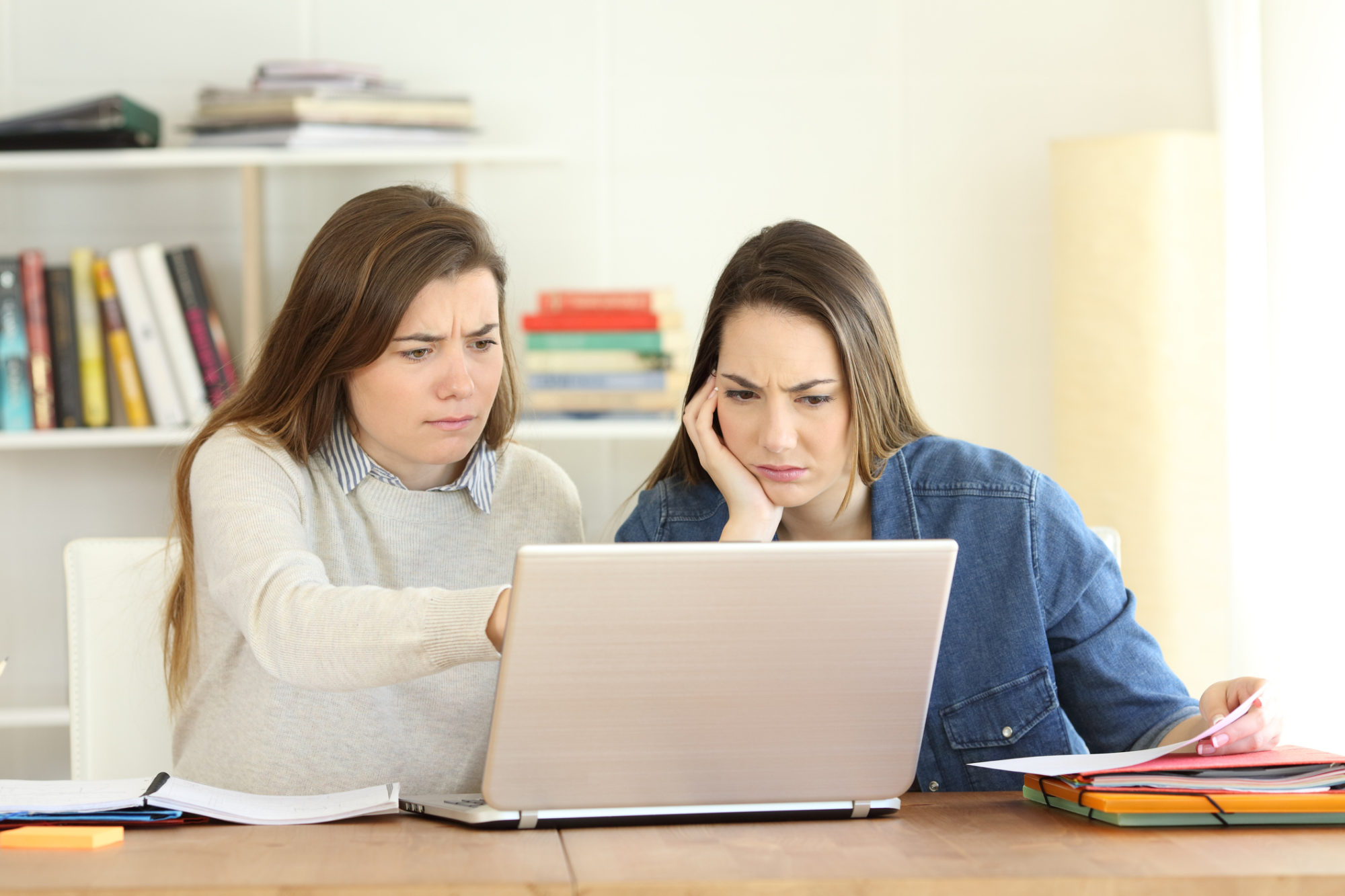 Two women looking at a laptop on a table at home with confused expressions.