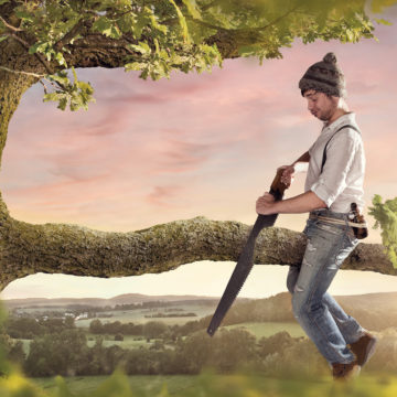 A man with a hat is sitting on a branch he is about to cut off.